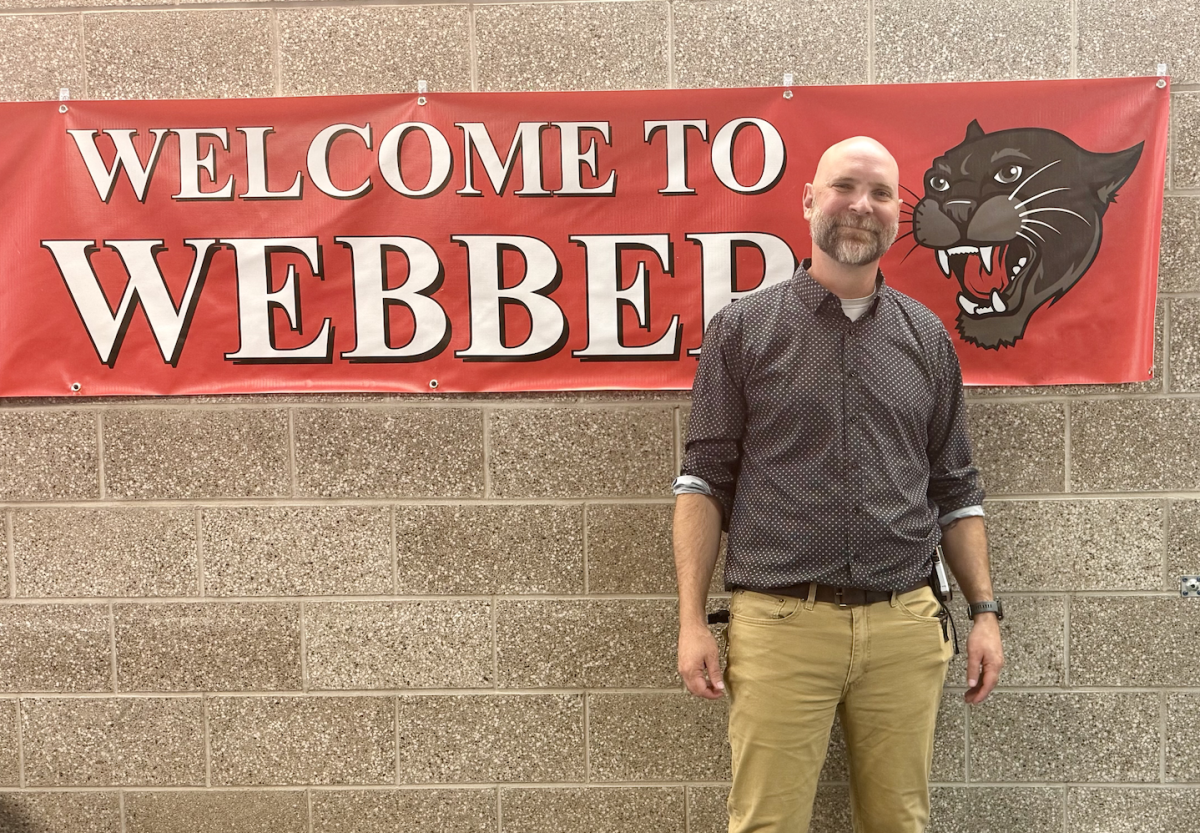 Mr. Steven Watkins, Webber's new principal, stands in front of a banner that says, "Welcome to Webber." The banner has the text on the left side with a panther picture on the right side.